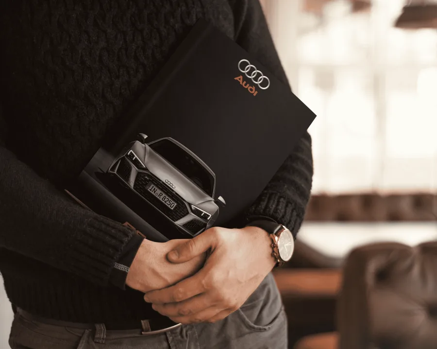 The Audi R8 catalogue held under an arm: a black cover with the Audi rings and a grey R8